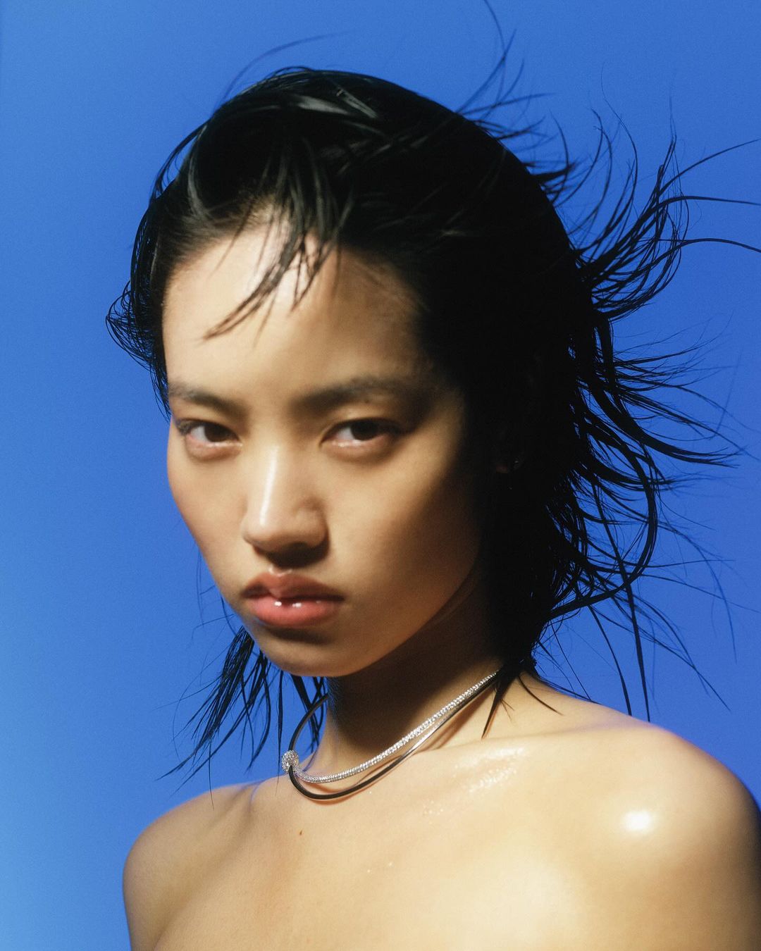Close-up portrait of a young Asian woman with wet black hair against a vibrant blue background, wearing a silver necklace.