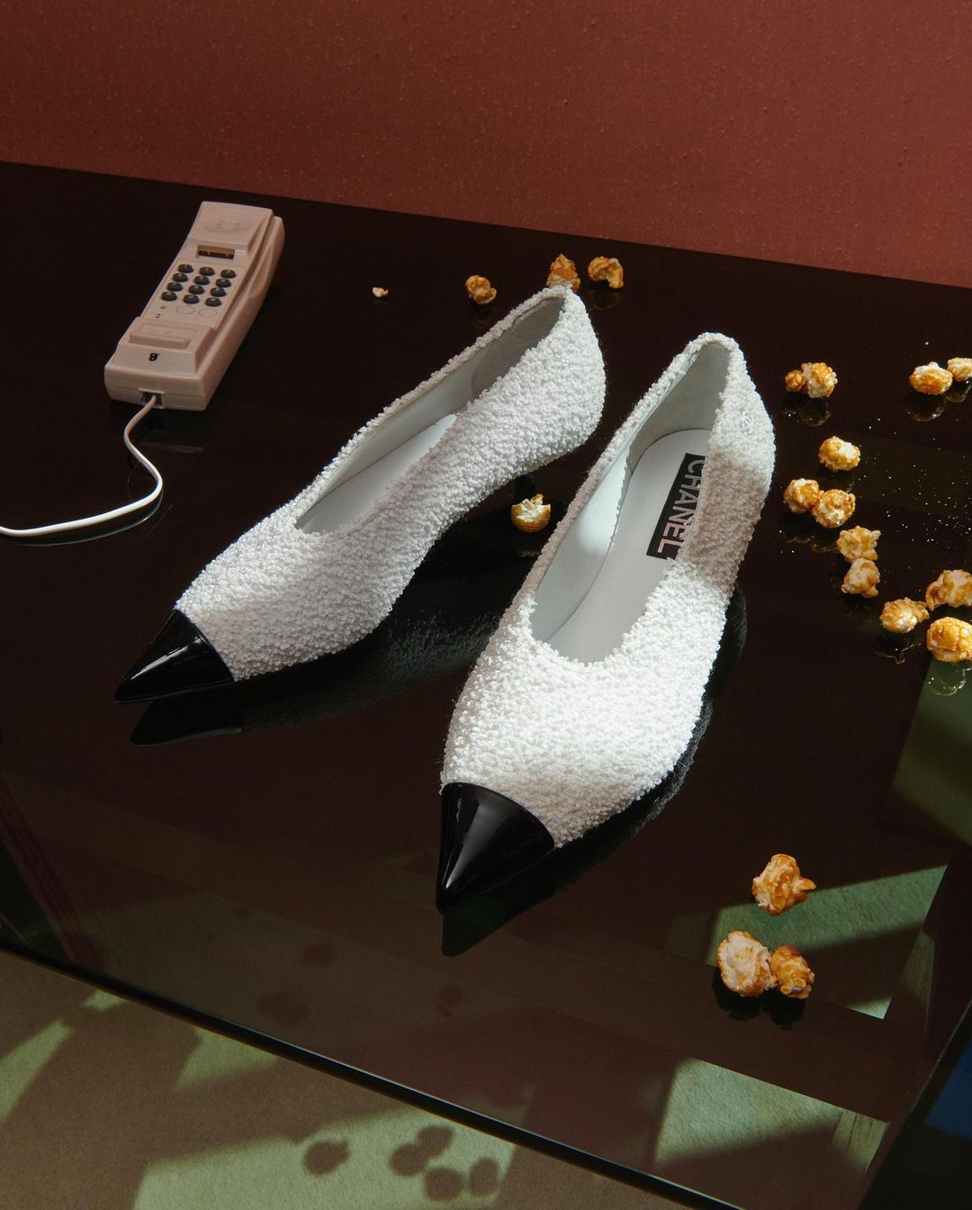 Elegant Chanel pumps in a still life with a vintage telephone and scattered popcorn on a reflective surface against a warm-toned wall.