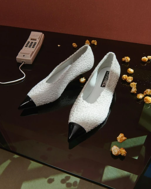 Elegant Chanel pumps in a still life with a vintage telephone and scattered popcorn on a reflective surface against a warm-toned wall.