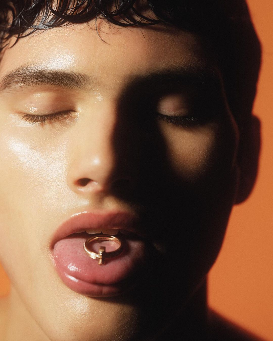 Close-up studio shot of a young adult with closed eyes, holding a gold ring on their tongue against an orange background.