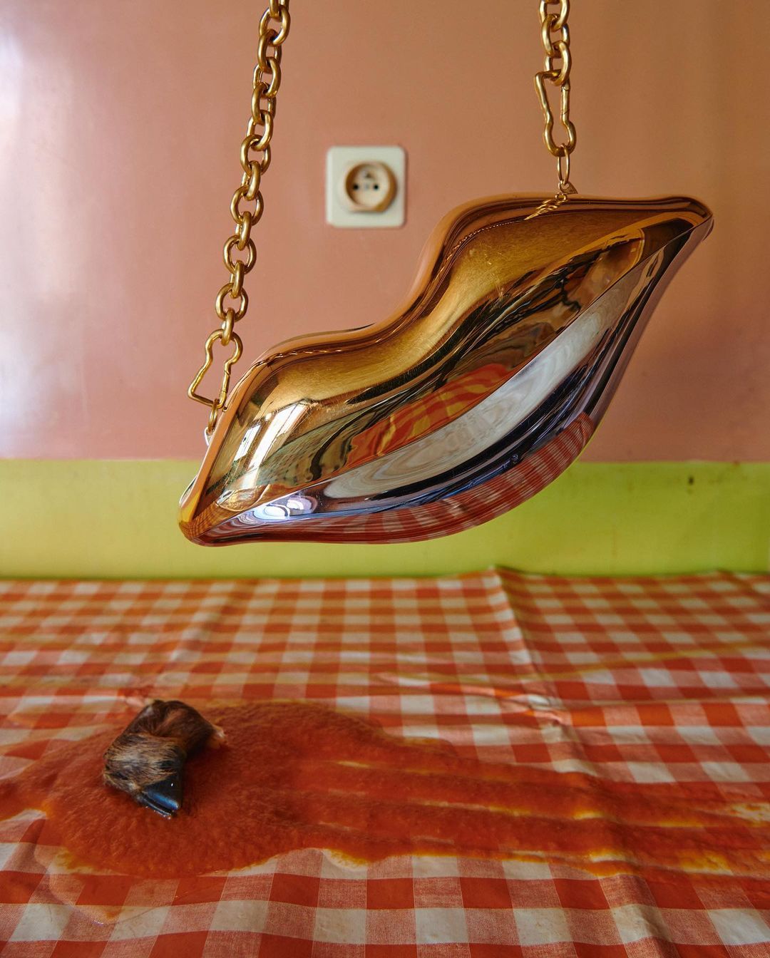 Gold lips-shaped purse hanging over a red and white checkered tablecloth with a spill on it, against a light pink and green wall.