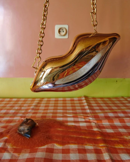 Gold lips-shaped purse hanging over a red and white checkered tablecloth with a spill on it, against a light pink and green wall.