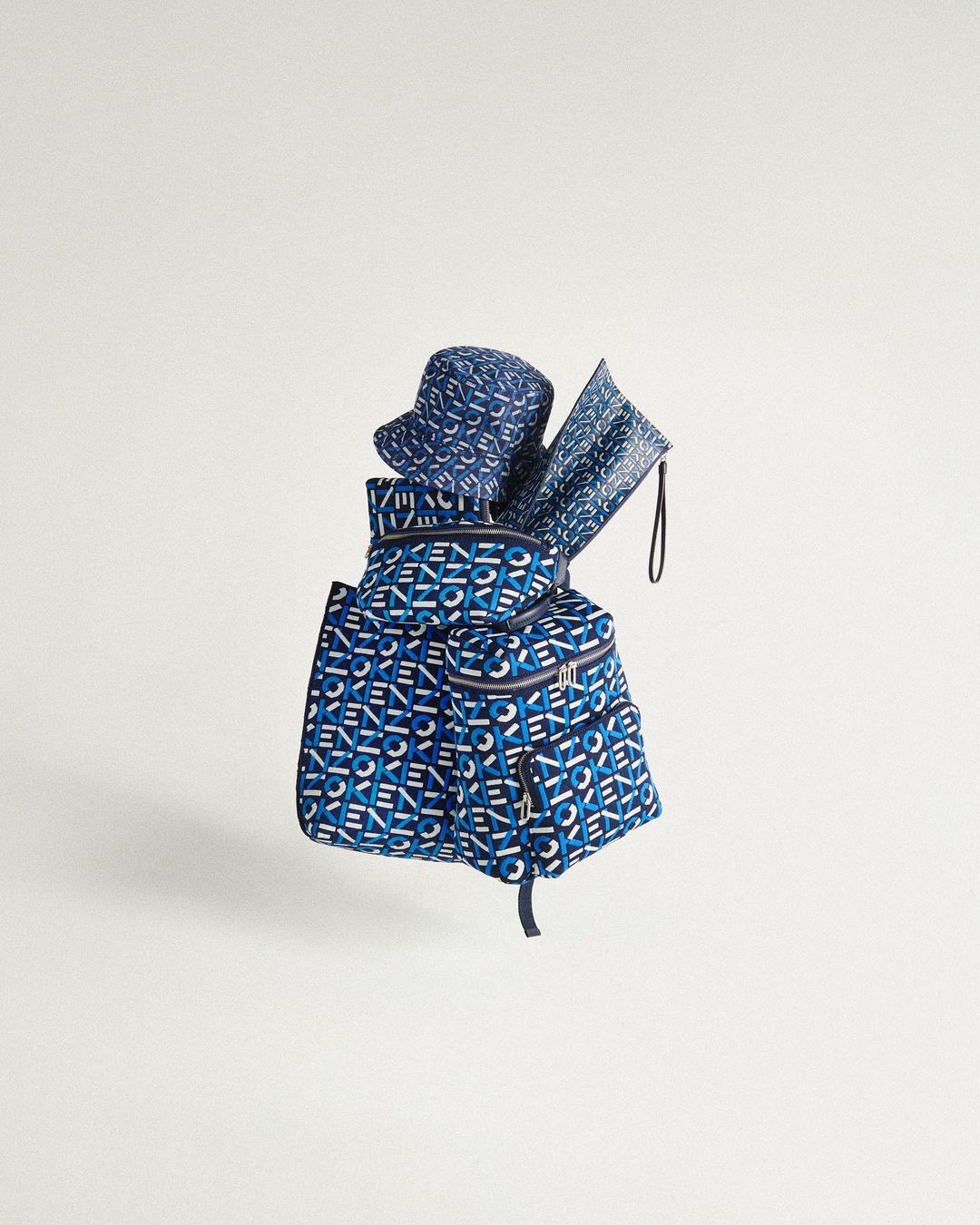 Studio shot of a blue backpack with the Kenzo logo pattern paired with a bucket hat and flat pouch, all against a clean white background.