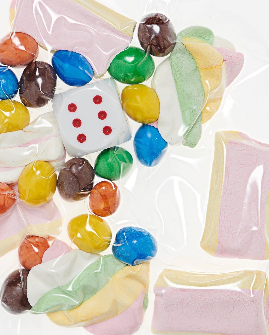 Colorful assortment of candy including chocolate balls, dice-shaped candy, and marshmallows in a clear packaging, studio lighting.