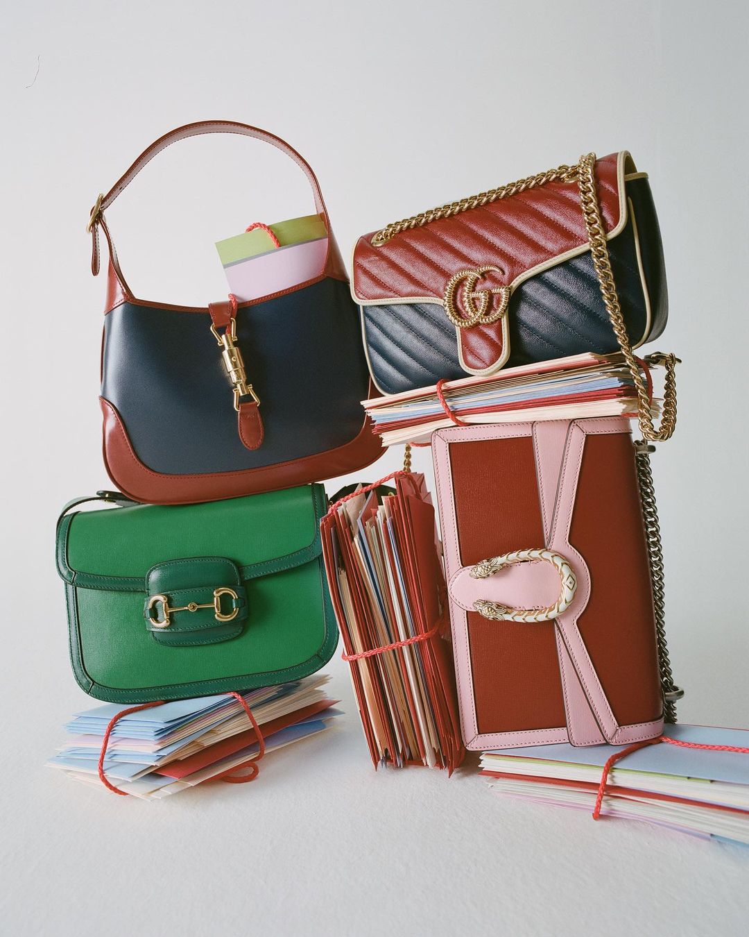 Stack of colorful Gucci purses and handbags, along with several bound stacks of paper, photographed in a brightly lit setting.