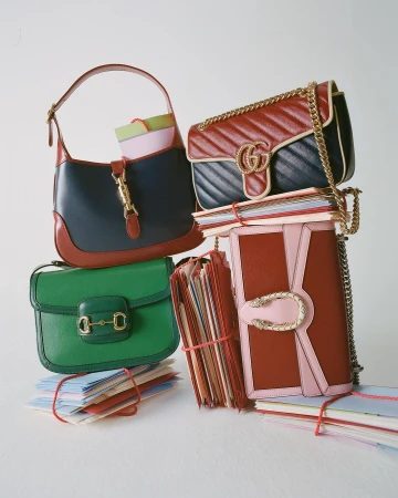 Stack of colorful Gucci purses and handbags, along with several bound stacks of paper, photographed in a brightly lit setting.