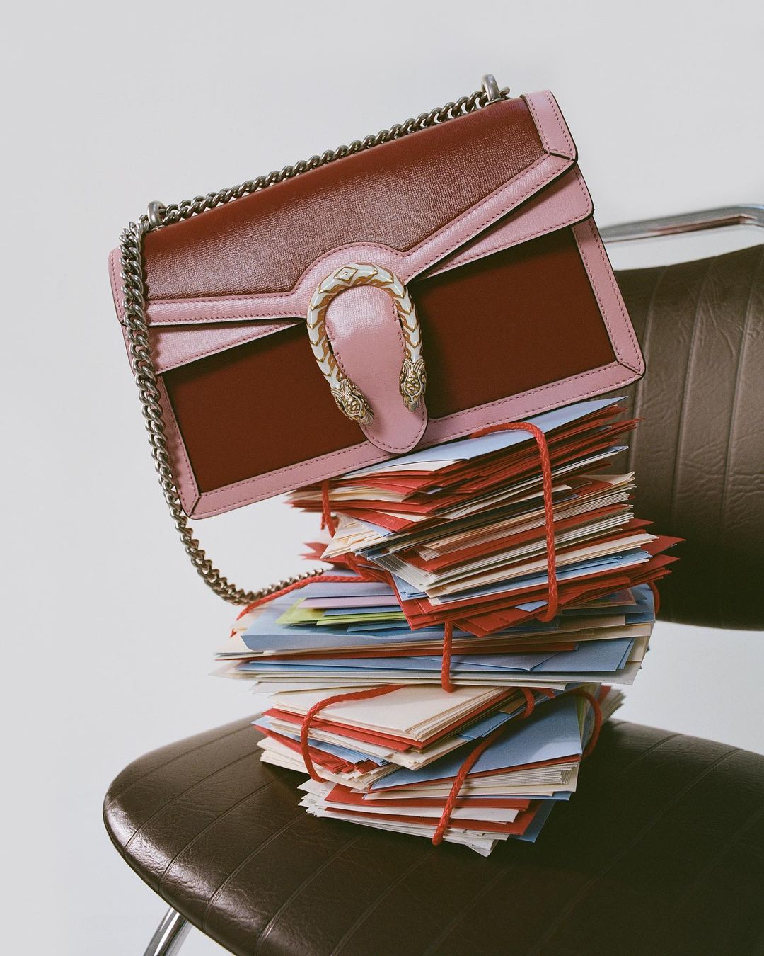 Burgundy and pink Gucci Dionysus bag displayed on a stack of cards tied with red string, set atop a brown leather chair.