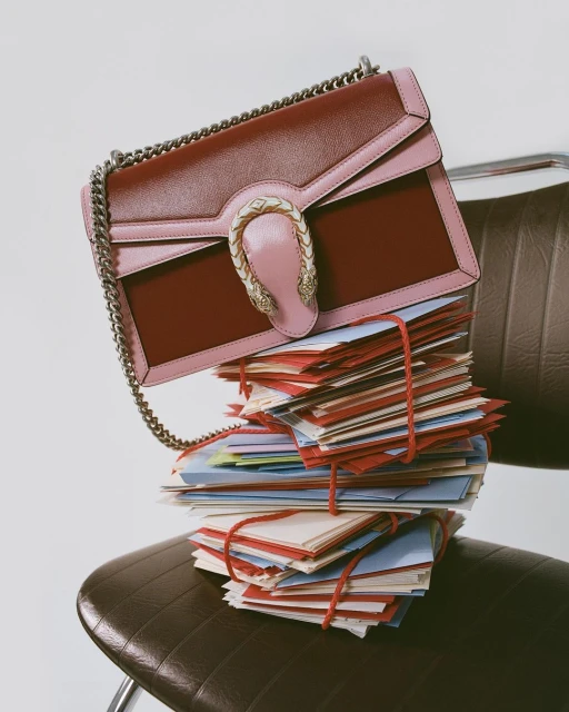 Burgundy and pink Gucci Dionysus bag displayed on a stack of cards tied with red string, set atop a brown leather chair.