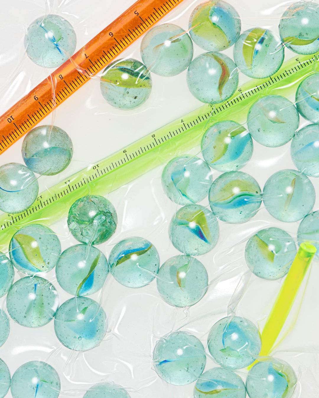 Overhead studio shot of clear marbles with blue and green swirls and multi-colored rulers in a clear plastic bag on white background.