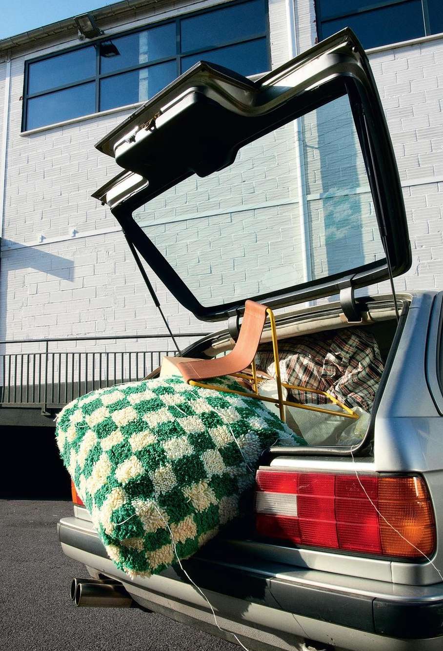 Loaded hatchback with checkered blanket and chair. Car parked near industrial building with open trunk showing objects inside.