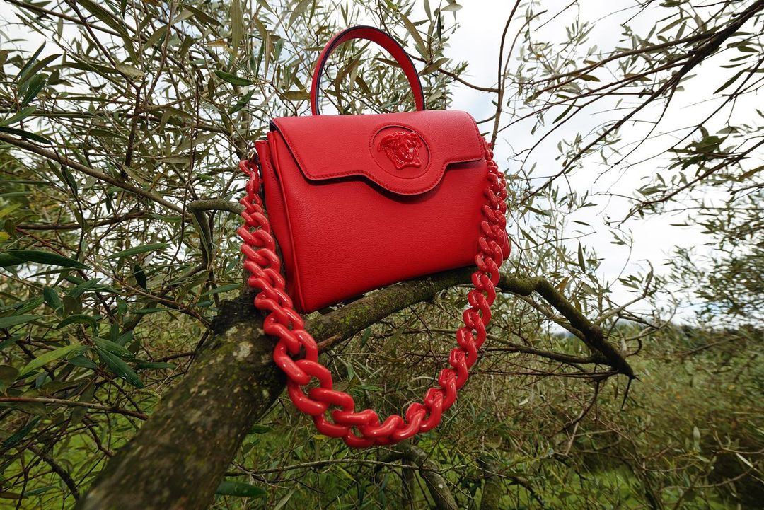 Bright red Versace purse with chain handle displayed on a tree branch outdoors, creating a striking contrast with the natural green foliage.