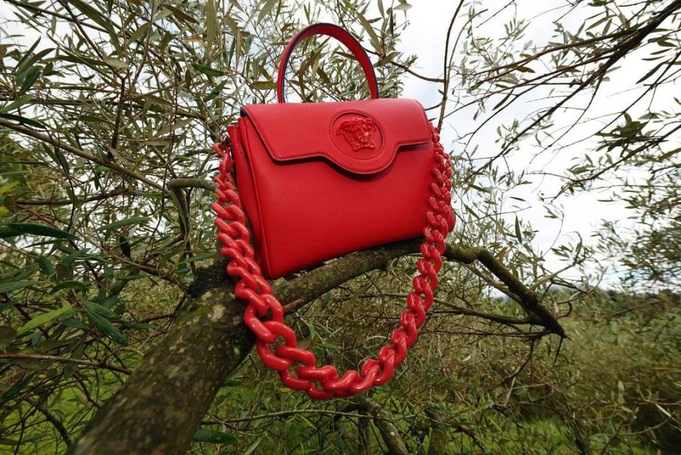 Bright red Versace purse with chain handle displayed on a tree branch outdoors, creating a striking contrast with the natural green foliage.