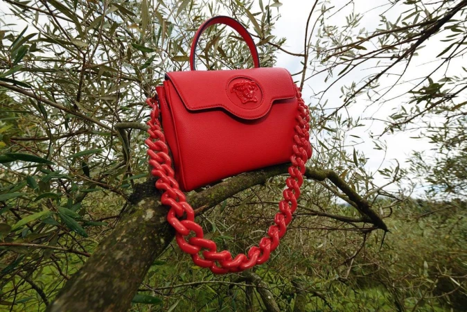 Bright red Versace purse with chain handle displayed on a tree branch outdoors, creating a striking contrast with the natural green foliage.