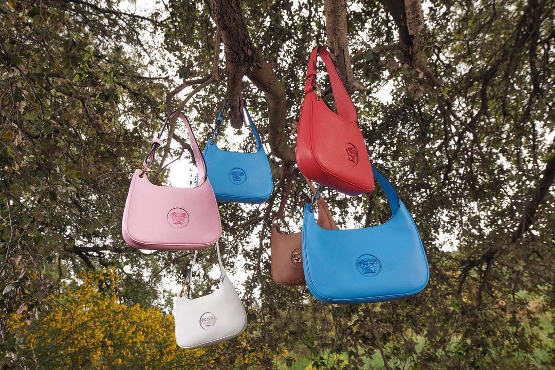 Versace handbags in various colors hang from tree branches in an outdoor fashion shot. The bags are pink, red, blue, brown and white.
