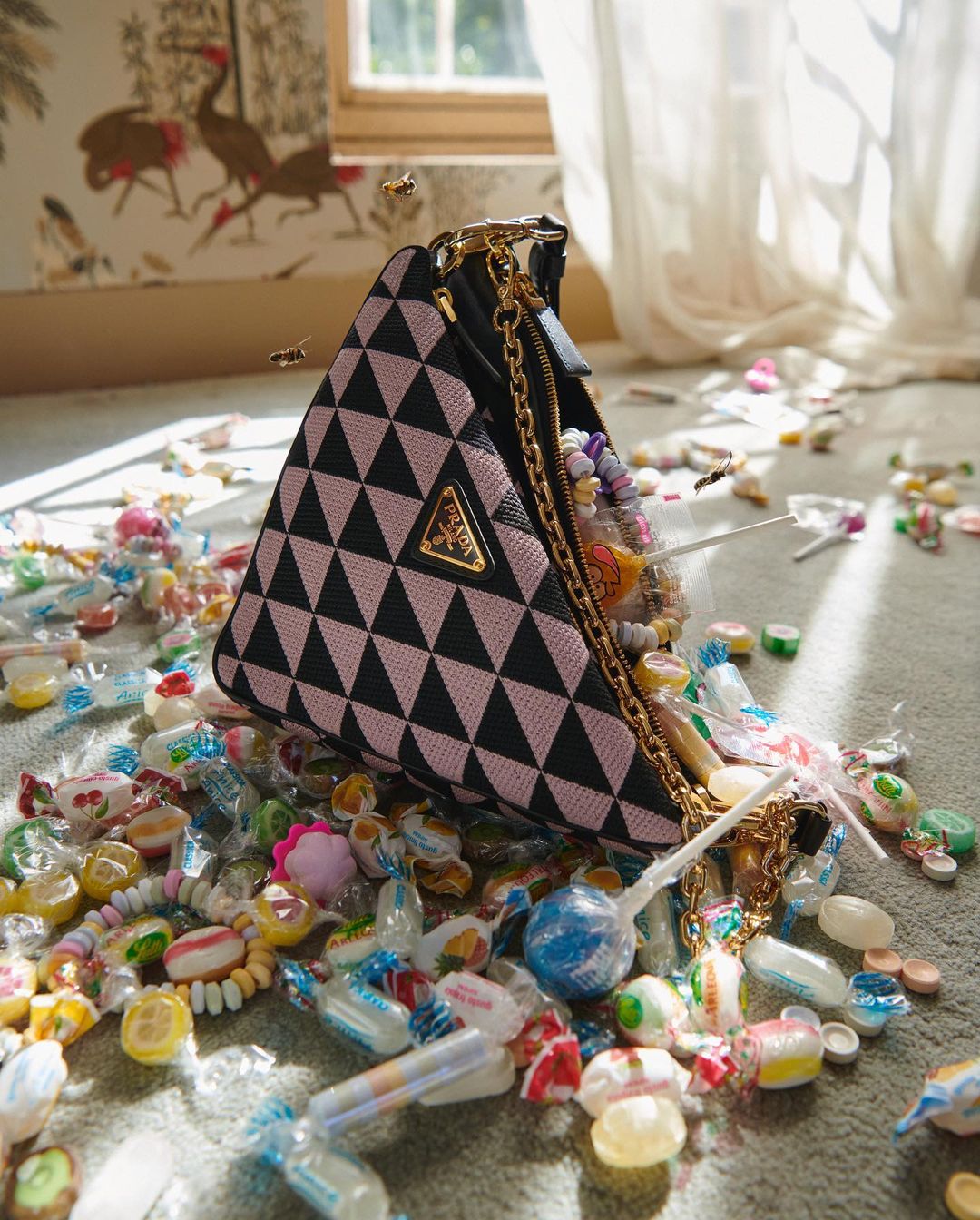 Designer handbag surrounded by assorted colorful candies in a brightly lit indoor setting with patterned wallpaper and natural lighting.