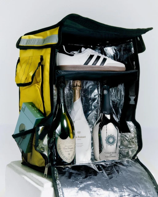 Open insulated delivery bag with champagne bottles, sneaker, and gift box inside. The bag is yellow with black trim and silver lining.