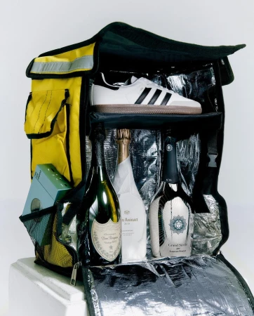 Open insulated delivery bag with champagne bottles, sneaker, and gift box inside. The bag is yellow with black trim and silver lining.