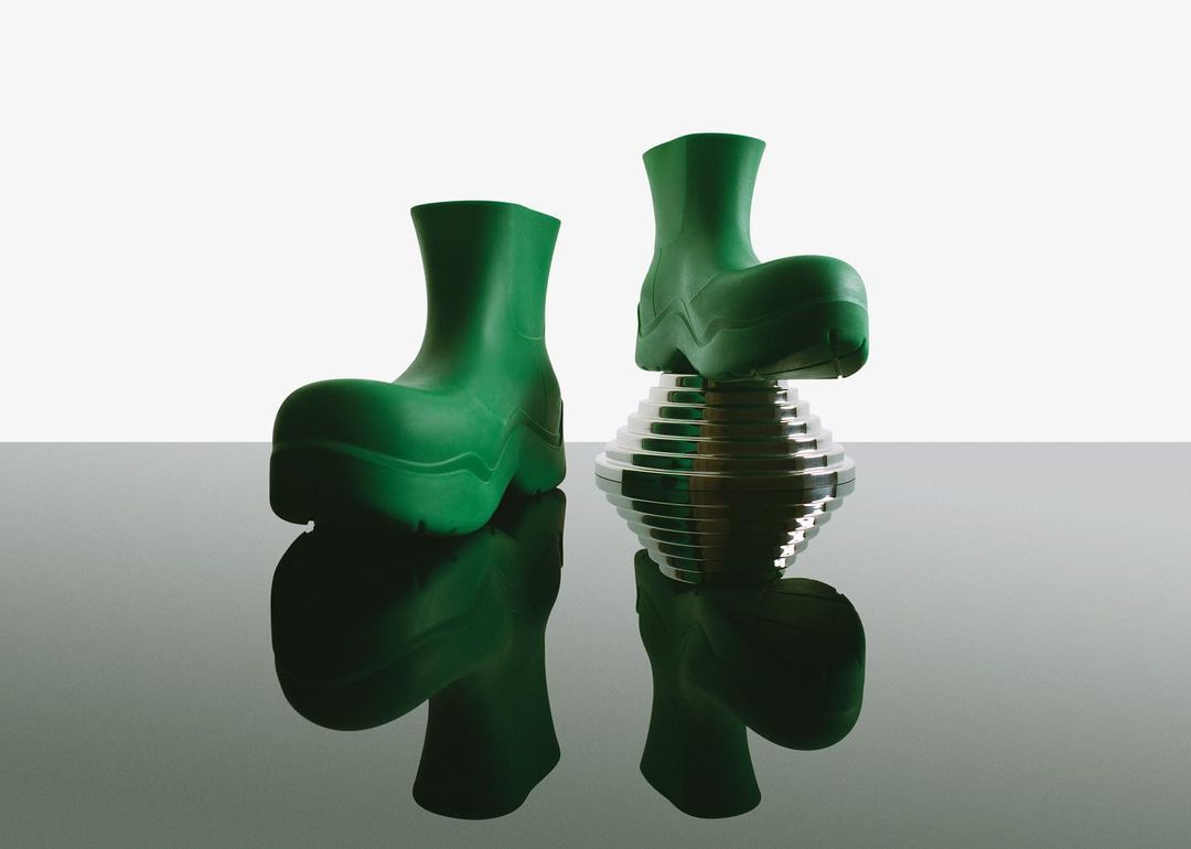 Pair of green rubber boots are displayed in a studio setting, one resting on a reflective silver platform for a modern footwear concept.