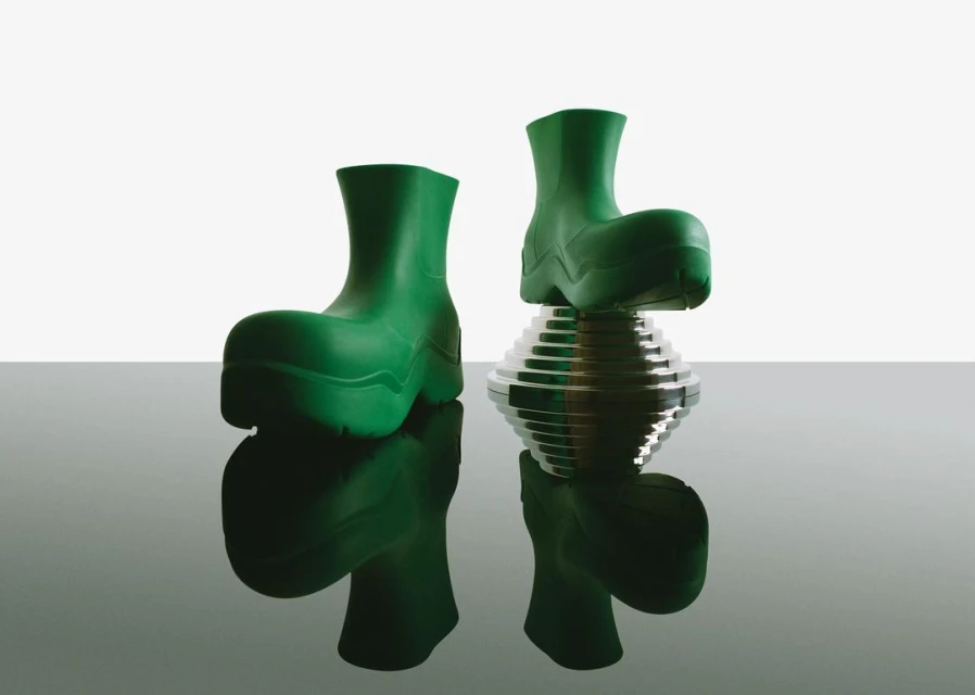 Pair of green rubber boots are displayed in a studio setting, one resting on a reflective silver platform for a modern footwear concept.