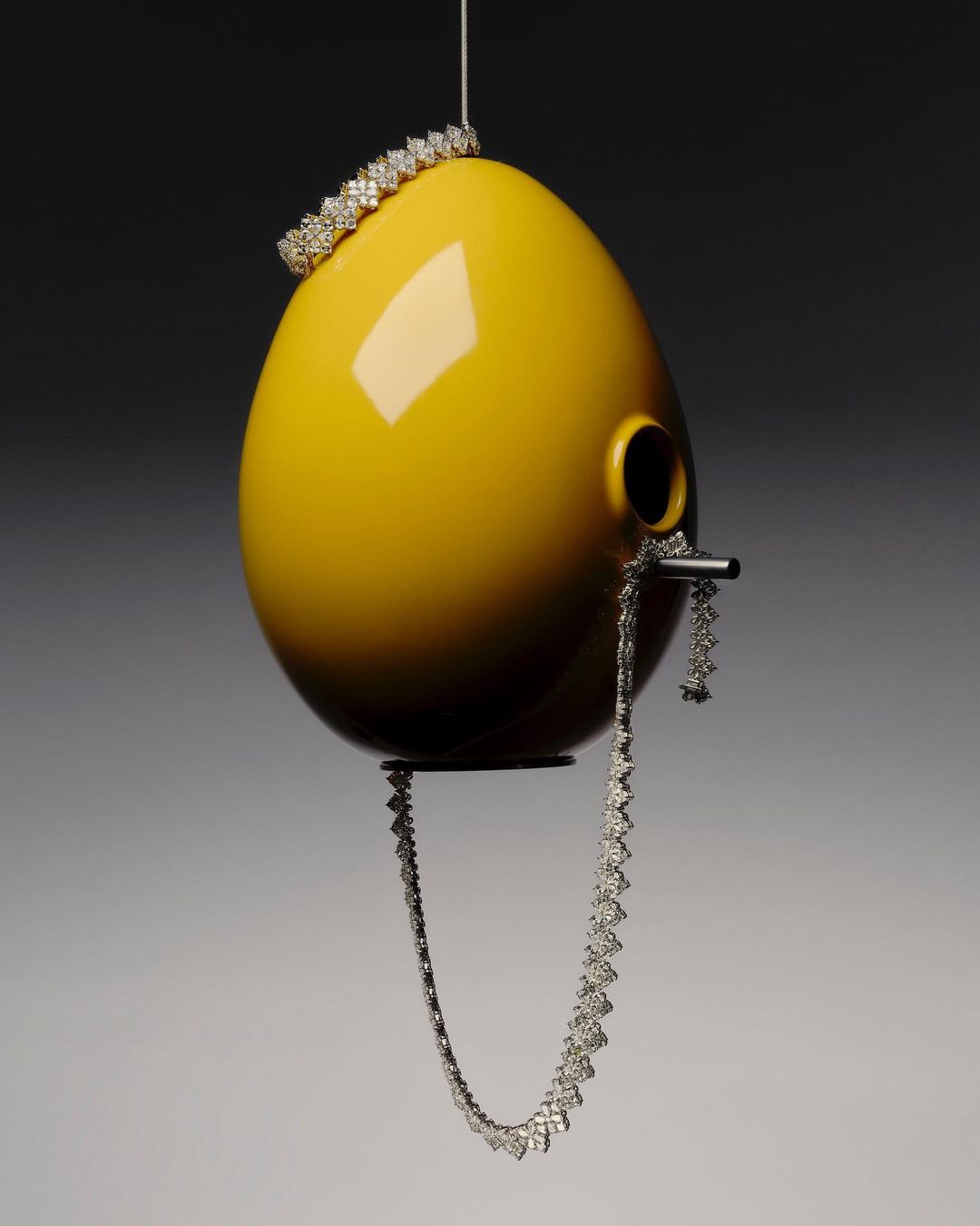 Yellow egg-shaped birdhouse with metal perch and jewelry accents including a silver tiara and necklace on a dark background.
