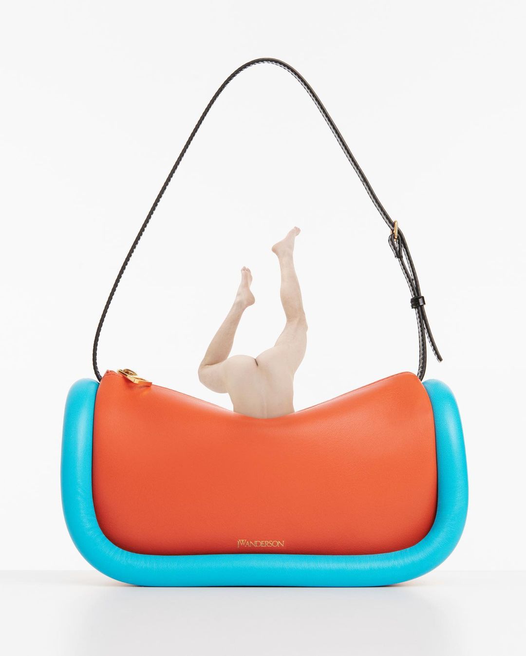 Bright orange and turquoise JW Anderson Bumper Moon handbag with black strap and disembodied human legs on a white backdrop.