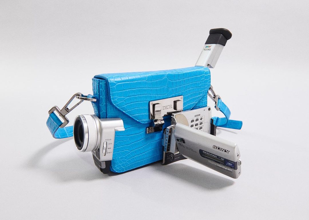 Novel handbag resembling a camcorder, featuring a bright blue, reptile-textured exterior and silver hardware against a plain white backdrop.