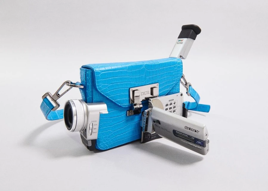 Novel handbag resembling a camcorder, featuring a bright blue, reptile-textured exterior and silver hardware against a plain white backdrop.