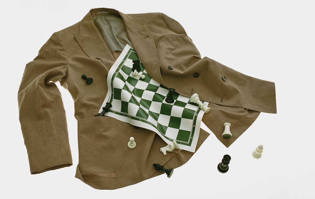 Wrinkled beige suit jacket with buttons, green and white checkered game board, and scattered black and white chess pieces on a white background.