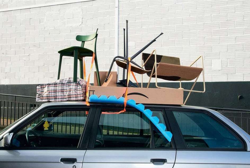 Silver station wagon overloaded with a green chair, tan chairs, boxes, and a blue pool noodle against a white brick wall.