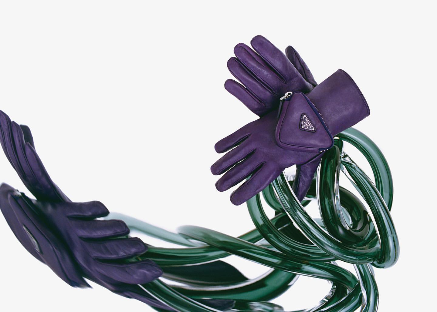 High-fashion still life featuring purple leather gloves with a Prada logo pocket, intertwined with translucent green, glossy tubing on white.
