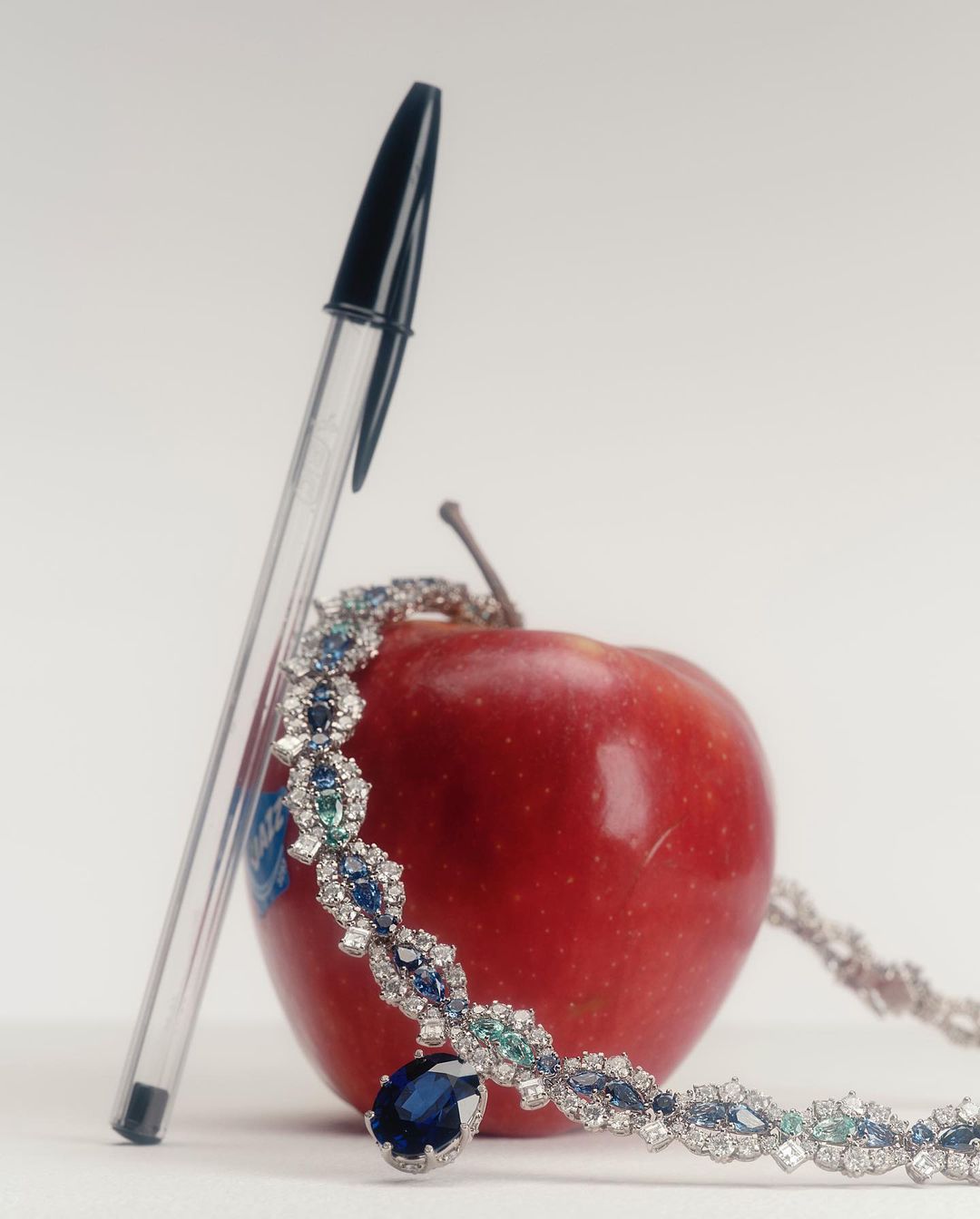 Elegant sapphire pendant necklace with diamonds, beside an apple and clear pen. Jewellery product shot for luxury fashion and design.