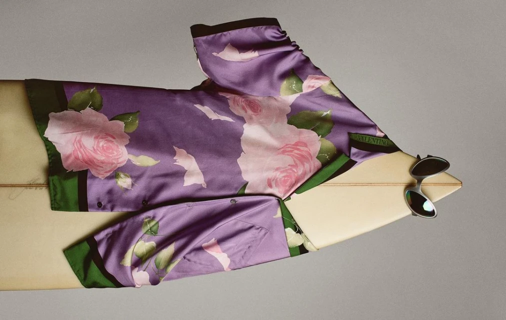 Floral purple button-down shirt draped across a surfboard with sunglasses. Fashion layout showcasing summer style.
