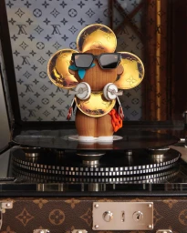 Wooden toy figure with gold monogram flower discs standing on a turntable inside a monogram trunk.