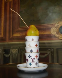 A ripe pear with a thin wooden stick pierced through the top, balanced vertically on a stacked tower of four patterned porcelain cups