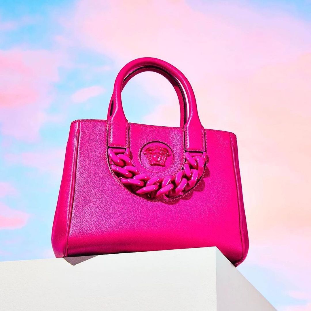 Close-up studio shot of a bright pink Versace handbag on a white block against a sky-like background with pink and blue hues.