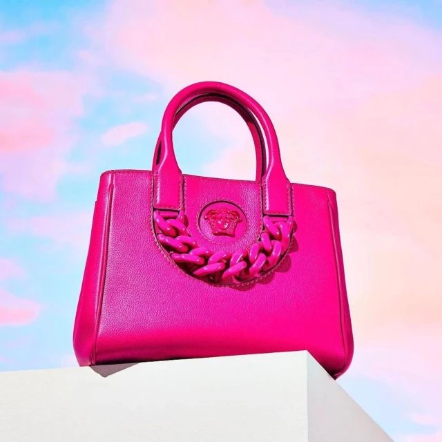 Close-up studio shot of a bright pink Versace handbag on a white block against a sky-like background with pink and blue hues.