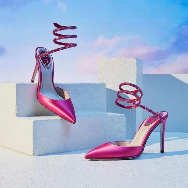 Two pink satin high-heel shoes are placed on white steps against a gradient blue and pink sky background in a fashion product shot.