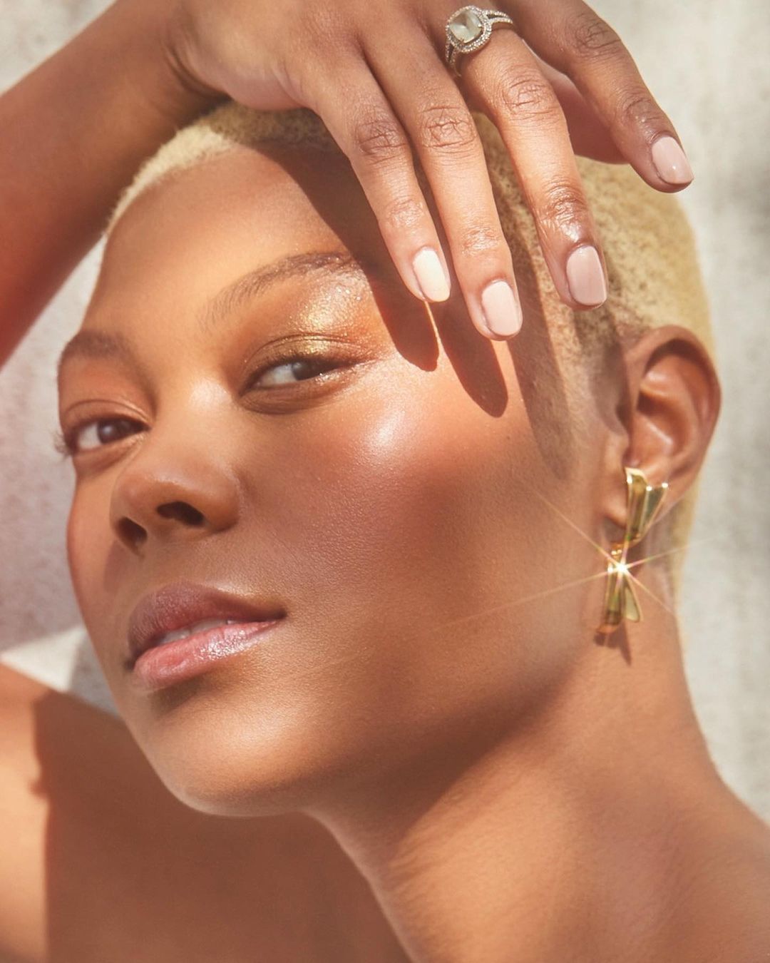 Close-up of a woman with a short blonde buzzcut and tan skin poses with a hand above her head showcasing a diamond ring and gold earrings.