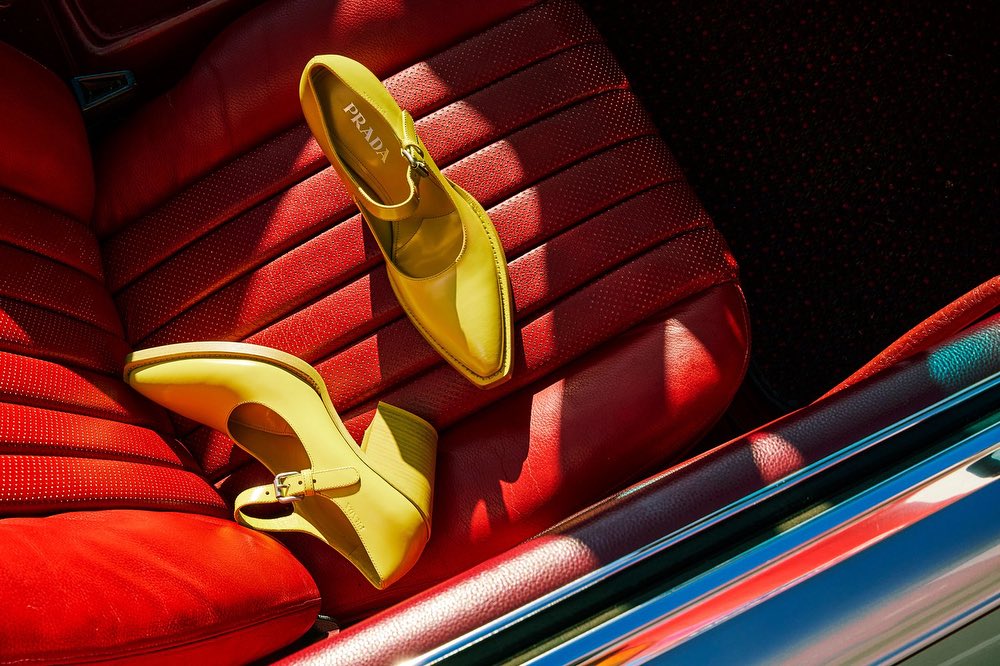 Pair of yellow Prada heeled shoes on a red leather car seat, showcasing a fashion accessory in a luxury automotive setting.