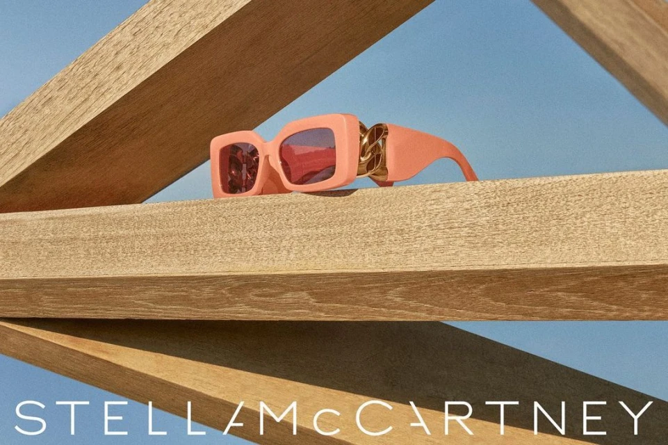 Peach-colored square sunglasses with gold chain detail displayed on wooden beams against a light blue sky. Stella McCartney logo visible.