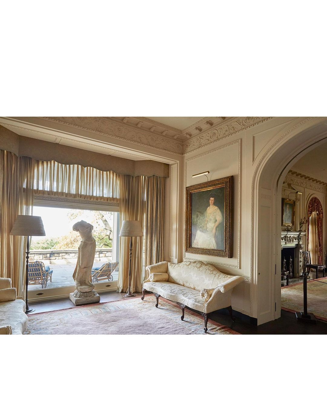 Ornate living room with a statue, painting, and lounge furniture, offering a glimpse of an outdoor patio, bathed in soft, natural light.