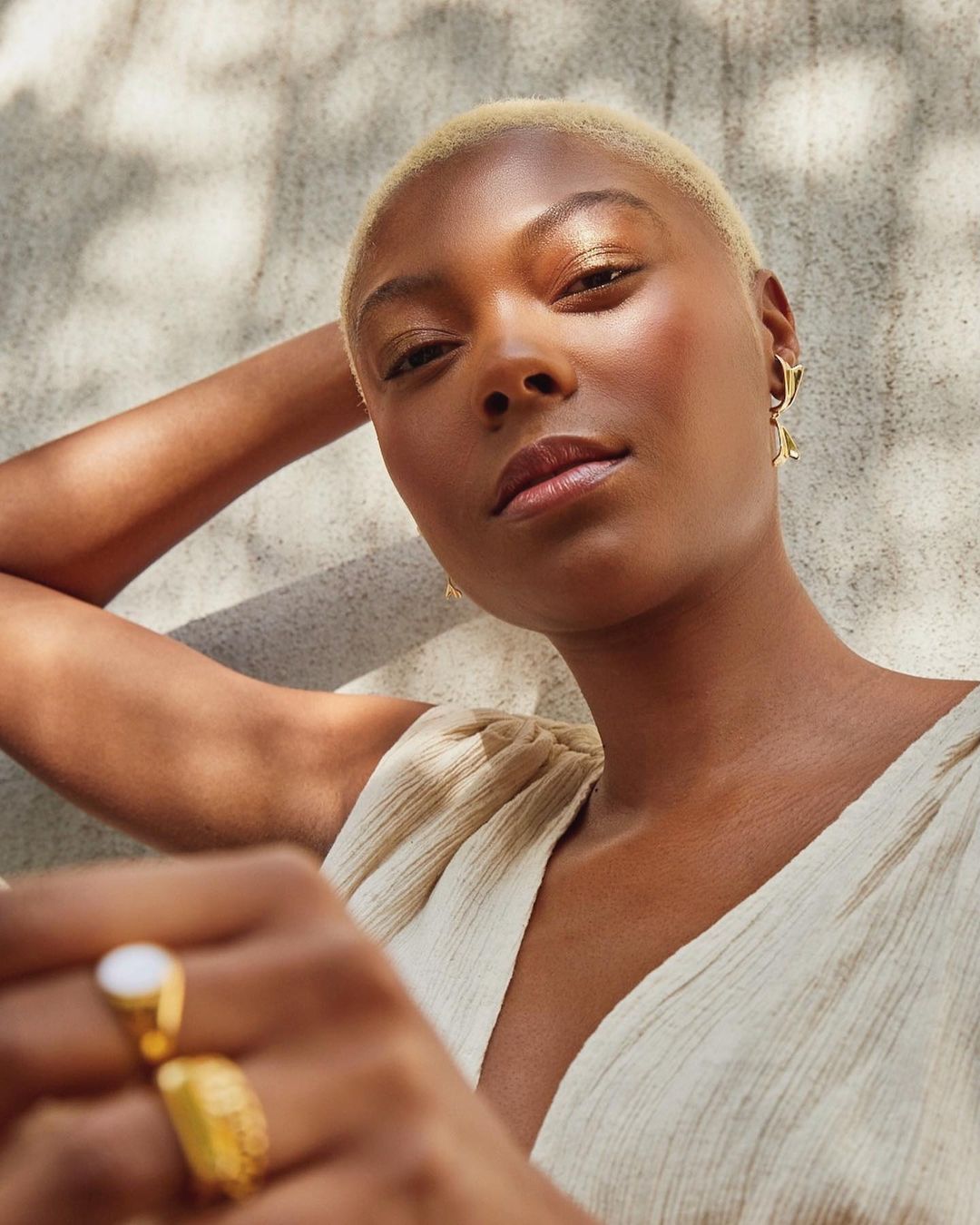 Close-up portrait of a young Black woman with short blonde hair wearing gold earrings and a ring, posing outdoors against a stone wall.