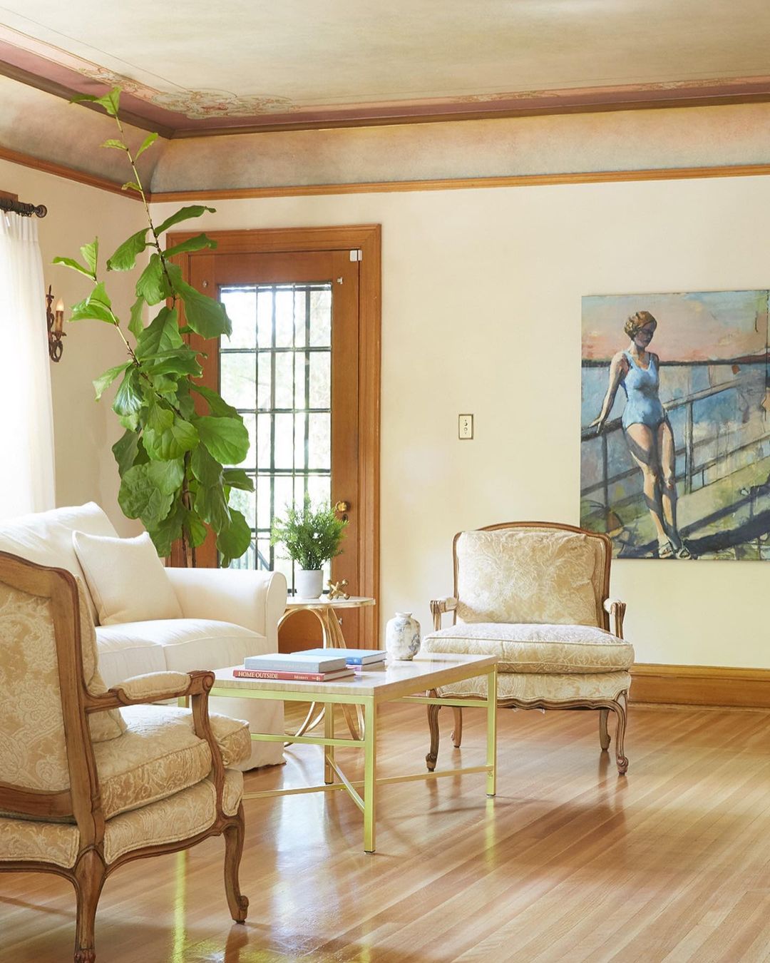Living room interior with two patterned armchairs, white sofa, coffee table, houseplant, and a painting of a woman in a swimsuit.