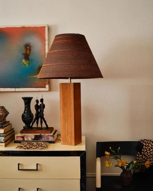 Interior still life featuring a wooden table lamp with a brown woven lampshade, decorative objects, and an abstract painting on the wall.