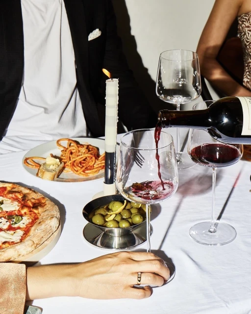 Red wine being poured into a glass at a table set with pizza, olives, and pasta, accompanied by a lit candle.