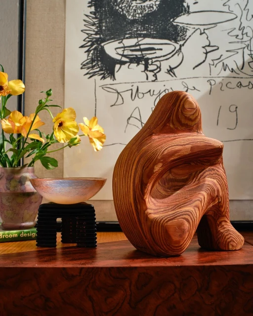Wooden abstract figure sculpture on a table with yellow flowers, a vase, and artwork. Warm lighting creates inviting mood.