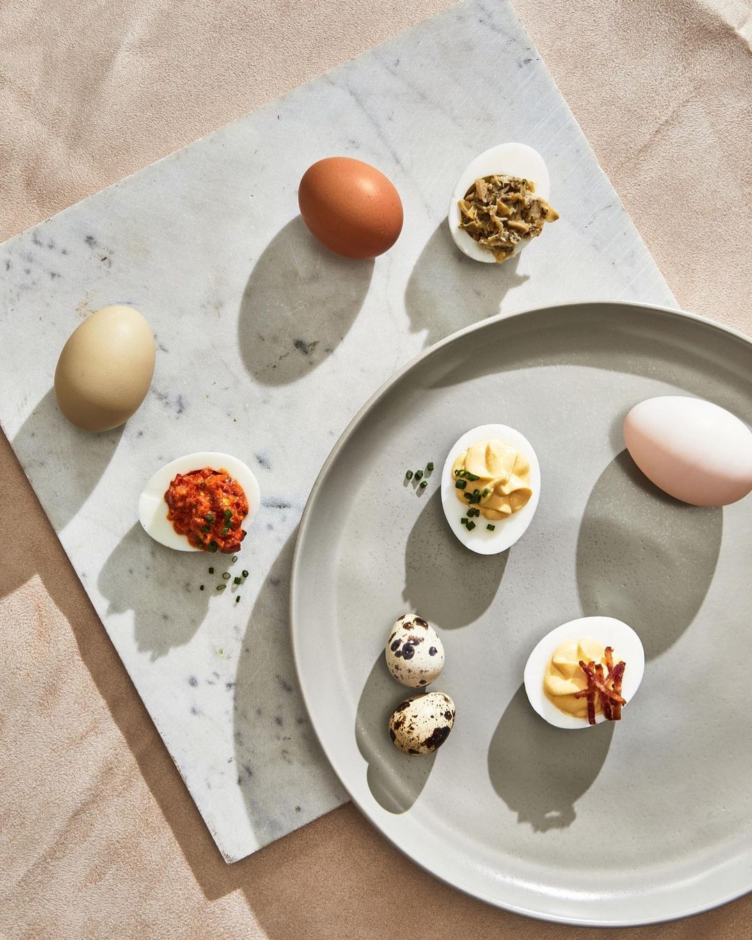 Overhead shot showcases a variety of egg dishes, including deviled eggs with various toppings, alongside whole eggs on a gray plate and marble board.