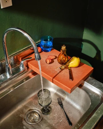 Still life in kitchen sink featuring kitchen utensils and a glass of water with light bulbs and cut tomatoes on an orange cutting board.
