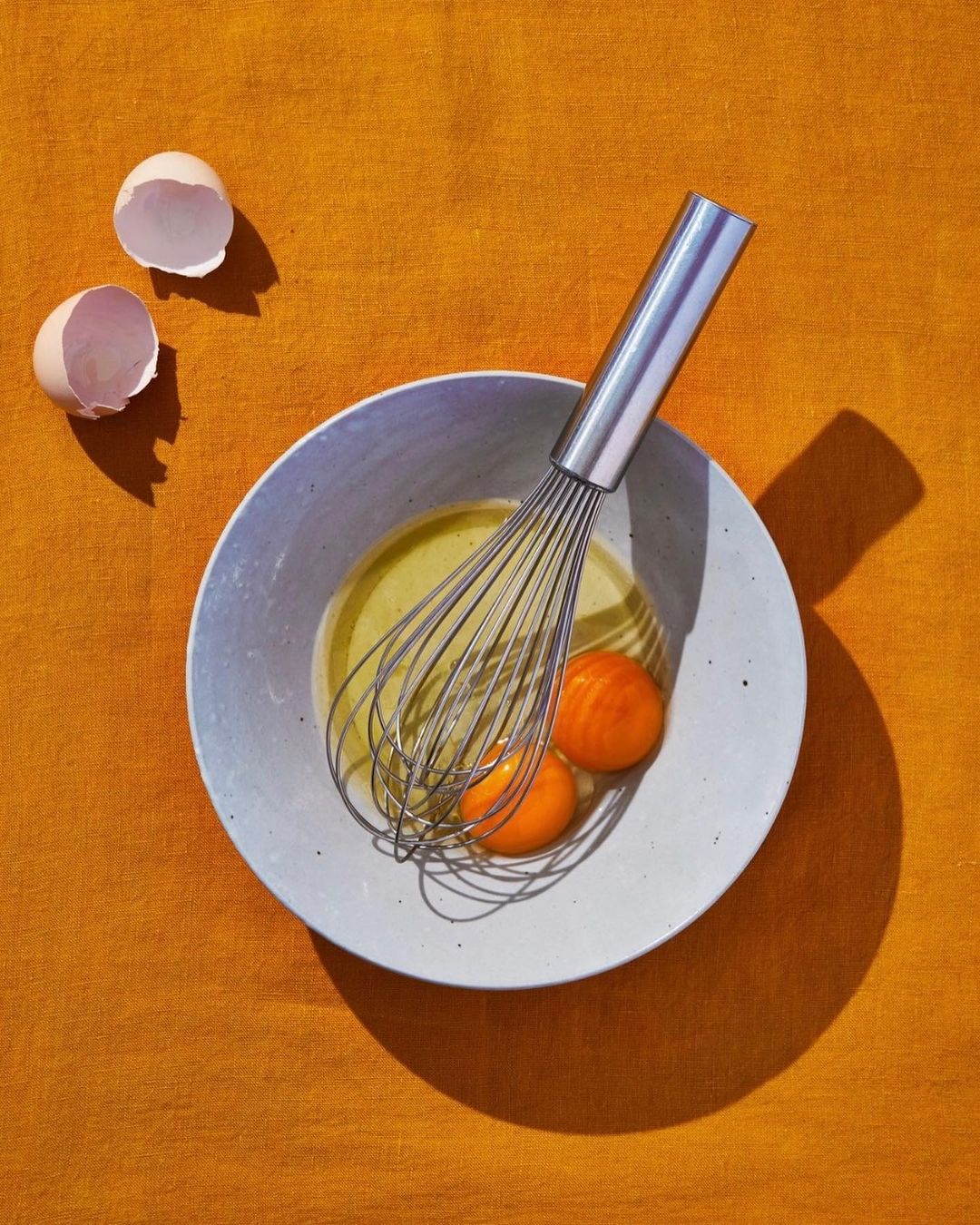 Overhead shot of a bowl with egg yolks and a whisk on an orange surface, alongside two eggshell halves.