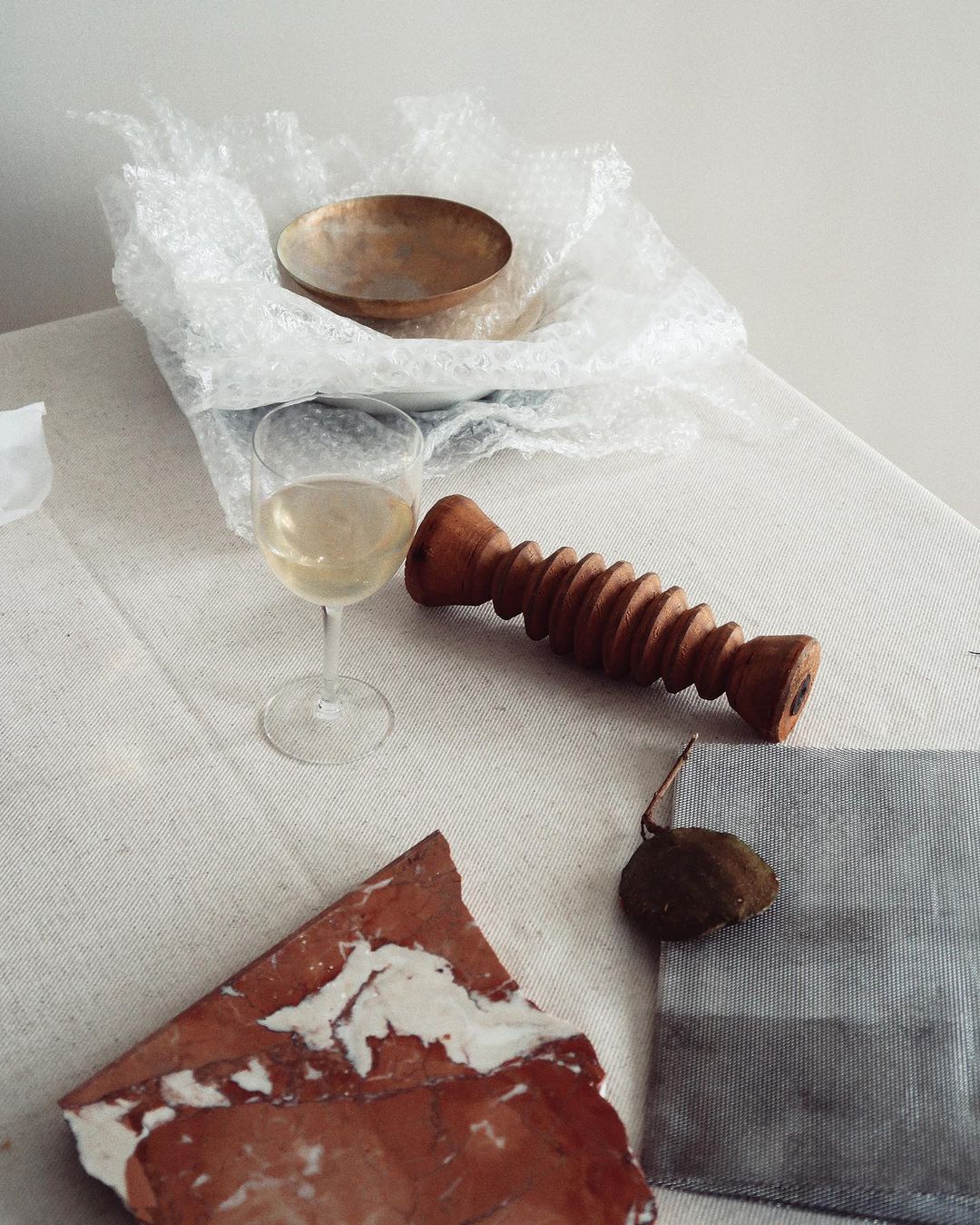 Still life of a bowl in bubble wrap, wine glass, wooden object, tile shard, cloth, and dried fruit, arranged on a white linen surface.